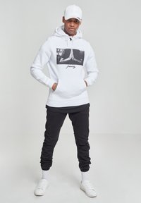 Hvit hettegenser med svart grafikk og teksten "pracy," kombinert med svarte joggebukser og hvite joggesko. Antrekket har en caps og er avslappet.