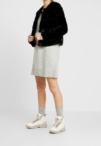 Kvinde iført en sort cropped faux fur jakke, grå strik sweaterkjole, grå sokker og hvide chunky snørebåndsstøvler, der står foran en ensfarvet baggrund.