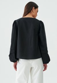 Top nero con scollatura quadrata, maniche a sbuffo e dettagli di tessuto stratificato nei polsini. Indossato con pantaloni bianchi. Texture liscia.