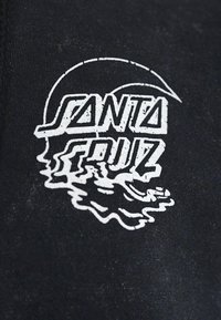 Svart stoff med hvit tekstlogo "SANTA CRUZ" med stilisert design, som inkorporerer flytende elementer og rund form.