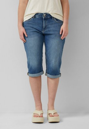 Personne portant un short en jean bleu retroussé aux genoux, un haut blanc et des sandales compensées beige, debout devant un fond gris.