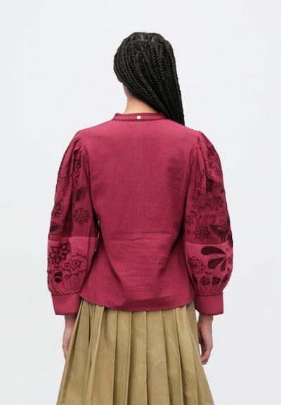 Tumši sarkana blūze ar apjomīgām izšūtām piedurknēm ar ziedu rakstiem. Gluda mugura ar apaļu kaklu un mazu pogas detaļu.