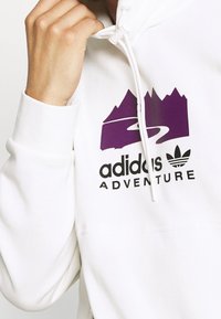 Vit hoodie med en lila bergsgrafik och texten "adidas ADVENTURE" i svart, samt en framficka och dragsko på huvan.