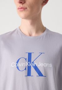 Light grey cotton t-shirt featuring a large blue "K" and "Calvin Klein Jeans" text in white. Round neckline, short sleeves, smooth texture.
