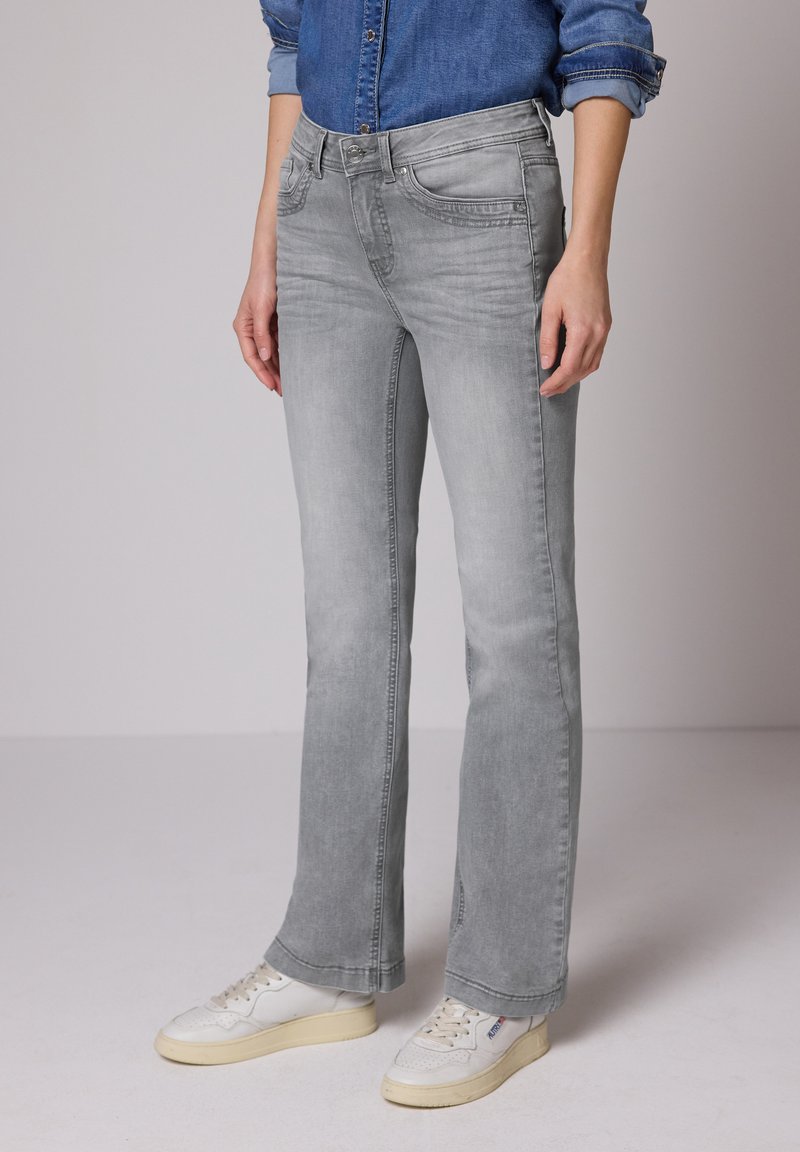 Personne portant un jean bootcut gris clair, des baskets blanches et une chemise en denim bleu aux manches retroussées, debout devant un fond uni.