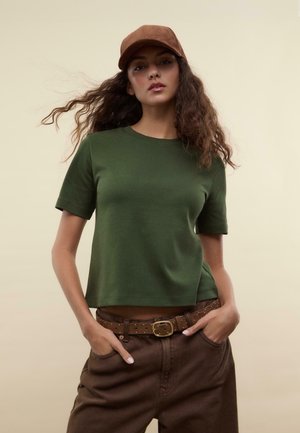 Groene katoenen t-shirt met korte mouwen, relaxte pasvorm, ronde halslijn, gecombineerd met bruine hoge taillebroek en een bruine pet.