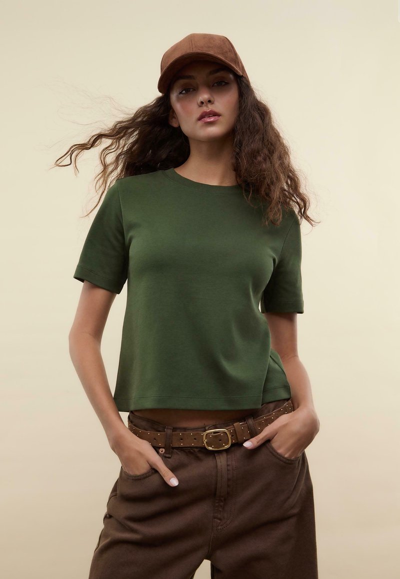 T-shirt verde in cotone a maniche corte, vestibilità comoda, scollatura rotonda, abbinato a pantaloni marroni a vita alta e un berretto marrone.