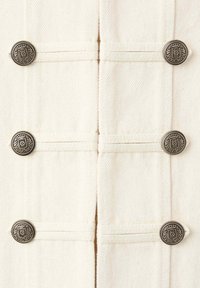 Six boutons décoratifs en argent disposés en deux colonnes verticales sur un tissu blanc texturé avec des bandes horizontales cousues.