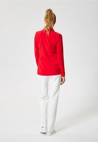 Chemise rouge à manches longues avec une coupe ajustée, poignets boutonnés, associée à un pantalon blanc et des chaussures claires. Texture lisse et lignes épurées.