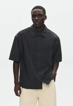 Mladić u teksturiranoj crnoj košulji s kratkim rukavima i džepom te bež hlačama, stoji ispred jednostavne bijele pozadine.