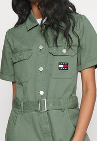 Grüner Baumwoll-Jumpsuit mit kurzen Ärmeln, zwei Brusttaschen, silbernen Knöpfen, einem taillierten Gürtel und einem Markenpatch auf der linken Tasche.