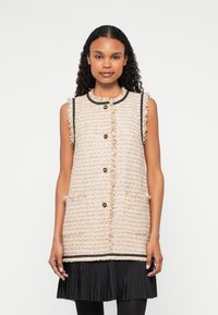 Beige tvídová bezrukávová vesta s kulat�ým límcem, třásněmi, černými detaily, zlatými knoflíky a bočními kapsami, přeložená přes černou skládací sukni.