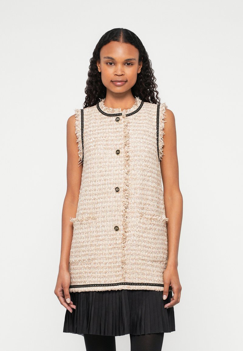 Beige tvídová bezrukávová vesta s kulatým límcem, třásněmi, černými detaily, zlatými knoflíky a bočními kapsami, přeložená přes černou skládací sukni.