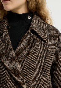 Bruin en zwart herringbone-patroon jas met een brede kraag, voorzien van een zilveren drukknoop en een zwarte geribde coltrui eronder.