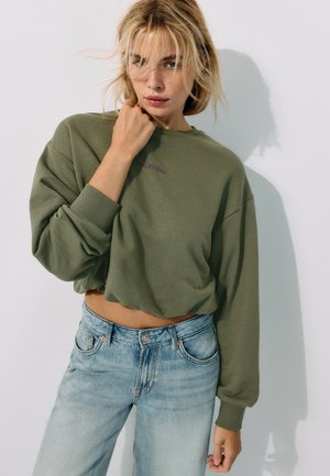 Lyshåret kvinde iført en olivengrøn cropped sweatshirt og lyseblå jeans, stående mod en plain hvid væg med hånden nær ansigtet.