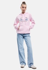 Różowa bluza z kapturem wykonana z miękkiego materiału, z graficznym tekstem i ilustracjami w stylu kreskówkowym. Połączona z jasnoniebieskimi spodniami z szerokimi nogawkami i casualowymi butami.