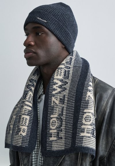 Mörkblå ribbad stickad mössa och mönstrad halsduk med växlande sektioner i marinblått och gräddvitt, med geometriska och bokstavsdesigns, bärs över en läderjacka.