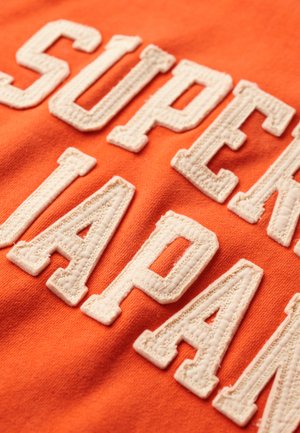 Orange bomuldsstof med store, broderede hvide bogstaver. Teksten lyder "SUPER JAPAN." Designet har en struktureret, hævet overflade.