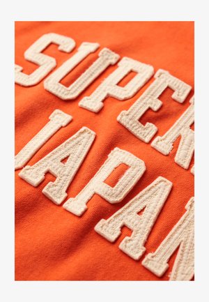 Orange bomuldsstof med store, broderede hvide bogstaver. Teksten lyder "SUPER JAPAN." Designet har en struktureret, hævet overflade.