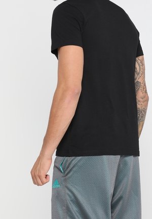 Man met getatoeëerde arm die een effen zwart T-shirt en grijze Adidas mesh shorts draagt, met zijn rug naar de camera.