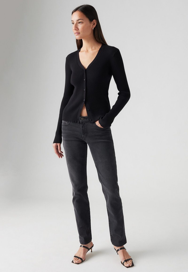 Cardigan noir côtelé avec un décolleté en V et des boutons, associé à un jean taille haute foncé et des talons noirs à brides. Textures lisses, design ajusté.