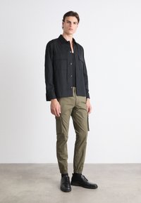 Camisa negra de botones con dos bolsillos en el pecho, combinada con pantalones cargo oliva que tienen bolsillos laterales y puños elásticos, usados con zapatos negros.