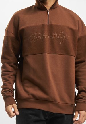 Man wearing brown quarter-zip sweatshirt with textured panel and cursive "Definitely" embroidery on the chest.