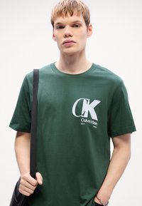 Zelena pamučna majica s istaknutim bijelim logotipom s natpisima "CK" i "Calvin Klein". Kratki rukavi, okrugli izrez, ležeran kroj.
