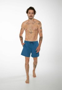 Modré plávacie šortky s elastickým pásom a šnúrkou, obsahujúce bočné vrecká a jednofarebný dizajn, nosené tetovanou osobou.