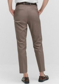 Osoba ubrana w beżowe, dopasowane spodnie i ciemnobrązowe mokasyny, stojąca z rękami w tylnych kieszeniach na jasnym, jednolitym tle.