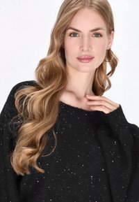Zwarte gebreide trui met een losse pasvorm, textuurafwerking met kleine zilveren accenten en brede mouwen. Het model heeft lang golvend haar en oorringen.