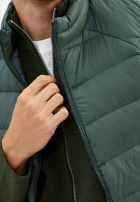 Ruka zapíná tmavě zelenou péřovku přes tmavě zelený svetr a bílou spodní košili.