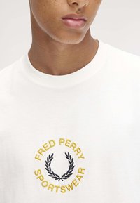 Camiseta de algodón blanca con escote redondo y un logotipo bordado en amarillo y negro, que representa una corona de laurel y texto.