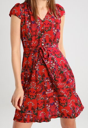 Frau trägt ein rotes, geblümtes Wickelkleid mit kurzen Ärmeln und einem gebundenen Taillengürtel.