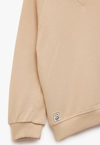 Sudadera de manga larga color beige con puños y dobladillo acanalados, que presenta un pequeño parche cuadrado con un logo de sol sonriente cerca del borde inferior.