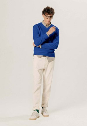 Jeune homme aux cheveux bouclés et lunettes, portant un pull bleu, une chemise à carreaux, un pantalon blanc, des chaussettes vertes et des baskets blanches, debout pensivement.