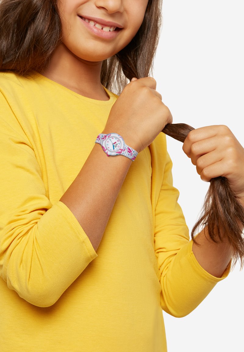 Børneur med pastelfarvet blå rem, der har pink og lilla mønstre. Rund skive med farverige markeringer, båret af et barn i en gul skjorte.