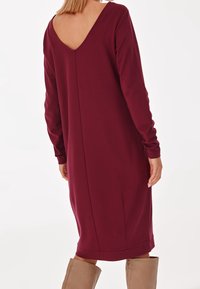 Burgundowa sukienka z długimi rękawami i głębokim dekoltem na plecach. Miękka, elastyczna tkanina; posiada szew na środku i zaokrąglony dół.