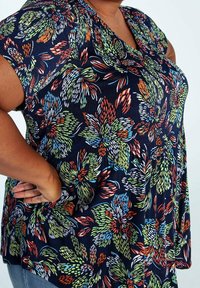 Femme portant une chemise bleu marine avec un motif floral abstrait multicolore, main posée sur la hanche, sur fond uni.