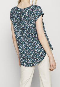 Blouse florale noire, avec un col rond, des manches courtes et un dos en forme de clé. Petits motifs de fleurs multicolores sur le tissu.