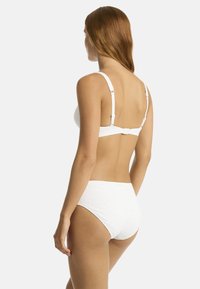 Frau mit langen roten Haaren, die einen weißen strukturierten Bikini trägt, von hinten gezeigt, vor einem schlichten hellgrauen Hintergrund.