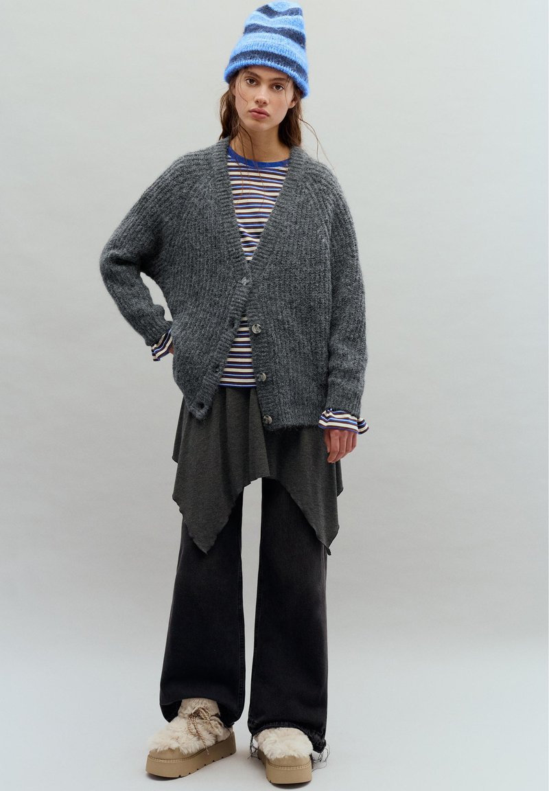 Jeune personne portant un bonnet tricoté rayé bleu, un cardigan gris oversize sur une chemise rayée, une jupe asymétrique, un pantalon large noir et des chaussures compensées doublées de fourrure.