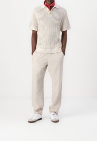 Polo de punto en color crema con una textura de tejido abierto, combinado con pantalones beige claros a medida. Zapatillas blancas con detalles en gris completan el conjunto.