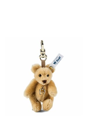 Pequeño llavero de peluche en forma de osito beige con cierre dorado, que lleva un colgante de herradura dorada y una etiqueta blanca de Steiff Collectors en la oreja.
