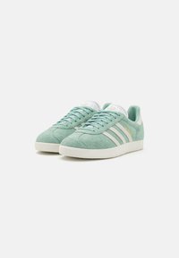 Zapatillas de ante verde con acentos en cuero blanco y tres rayas blancas. Punta redondeada, suela de goma plana y detalles en azul claro en el talón.