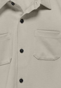 Chemise beige à boutons avec deux poches avant et boutons noirs, présentée en gros plan sur le tissu et les coutures.