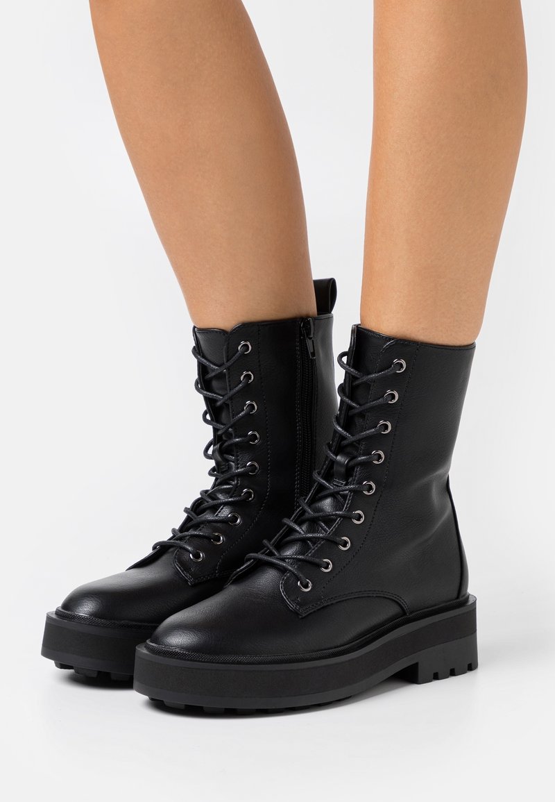 Bottes de combat en cuir noir à lacets avec semelles épaisses et épaisses portées sur des jambes nues, vues de face et de côté sur un fond blanc.