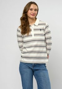 Jonge vrouw met lang golvend haar, gekleed in een gestreept shirt met lange mouwen en een blauwe spijkerbroek, staand tegen een effen achtergrond.