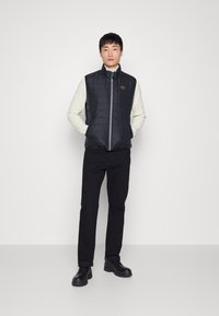 Sort quilted vest med høj krave, lynlås foran og et cirkulært logo patch, iført over en cremefarvet sweater, parret med sorte bukser og sko.