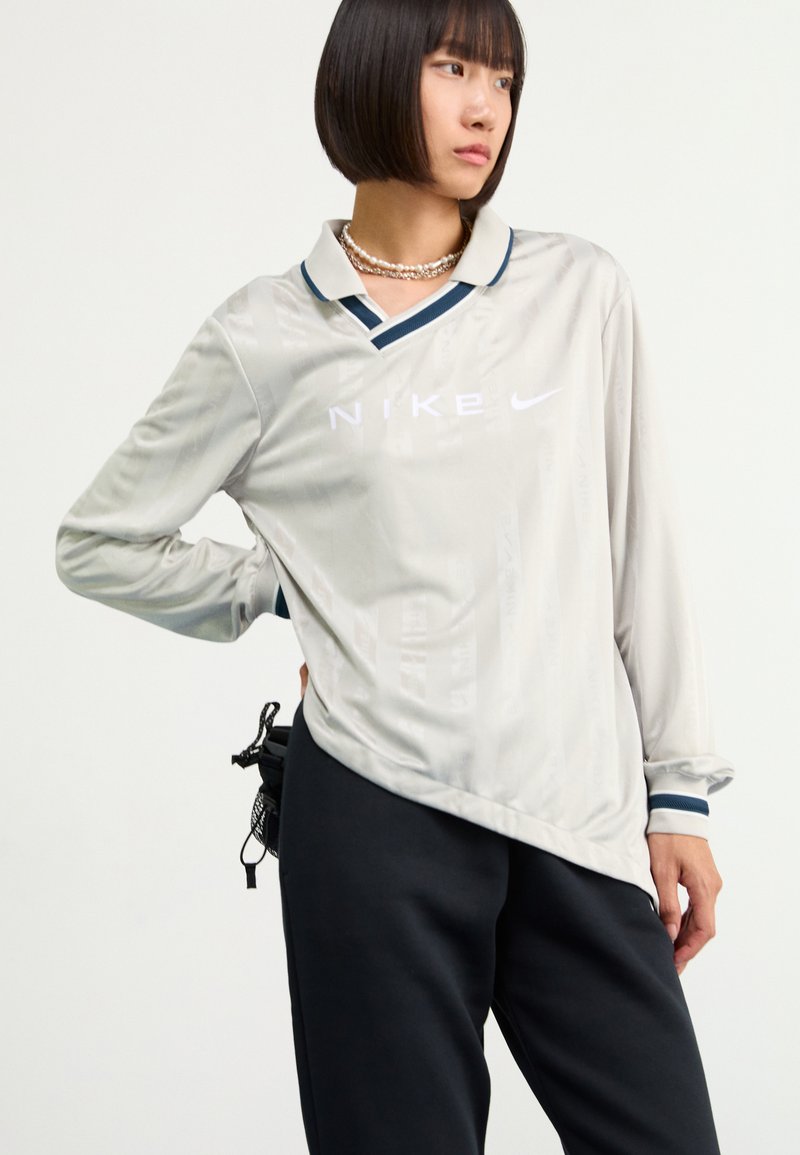 Chemise Nike à manches longues gris clair avec un col en V, arborant un logo blanc. Col et poignets en contraste bleu marine. Associée à un pantalon sombre.